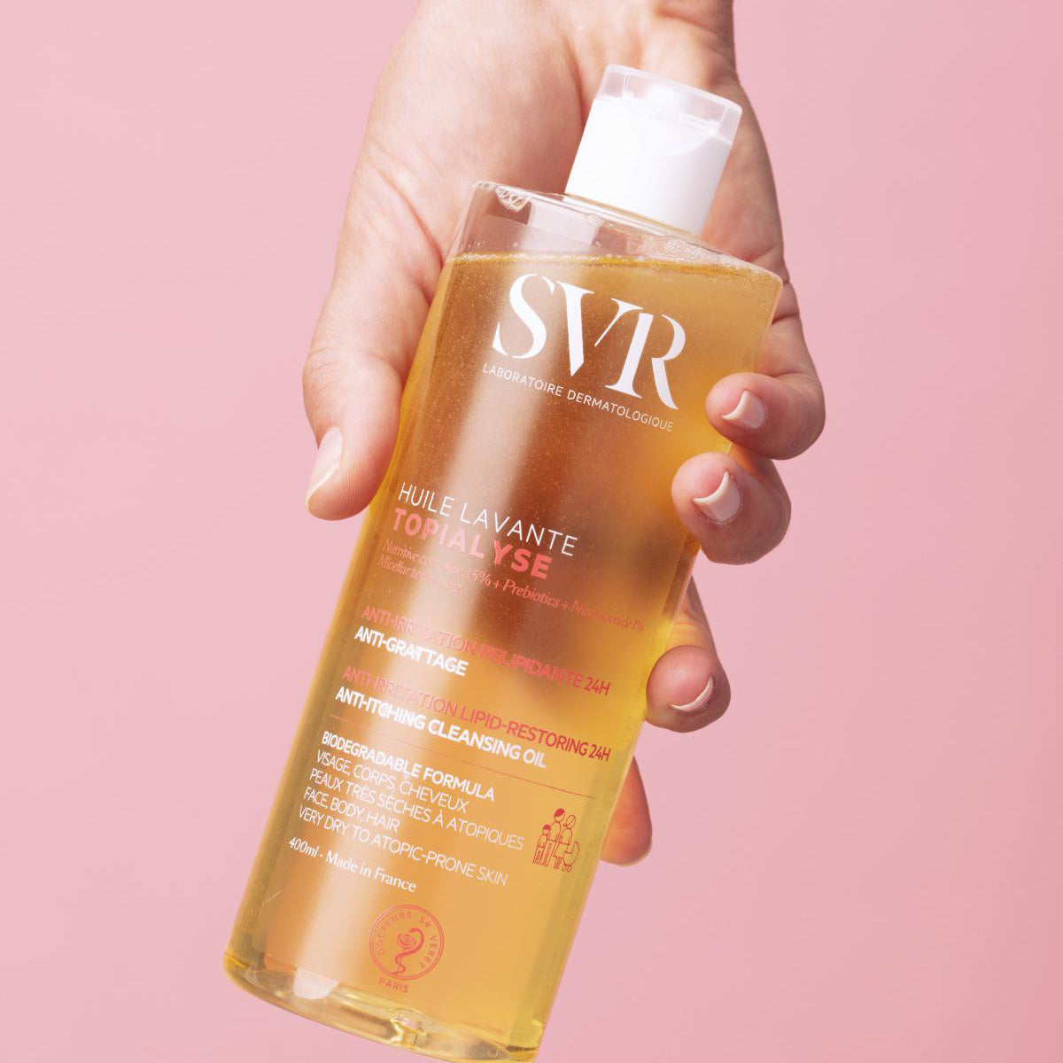 Main tenue par une main, flacon transparent de 400ml d'huile lavante SVR Topialyse à la texture dorée sur fond rose.