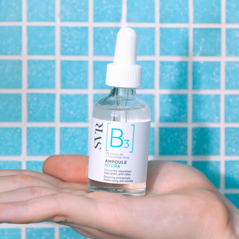 Flacon en verre de sérum SVR B3 Ampoule Hydra avec pipette blanche, tenu dans une main devant un carrelage bleu.