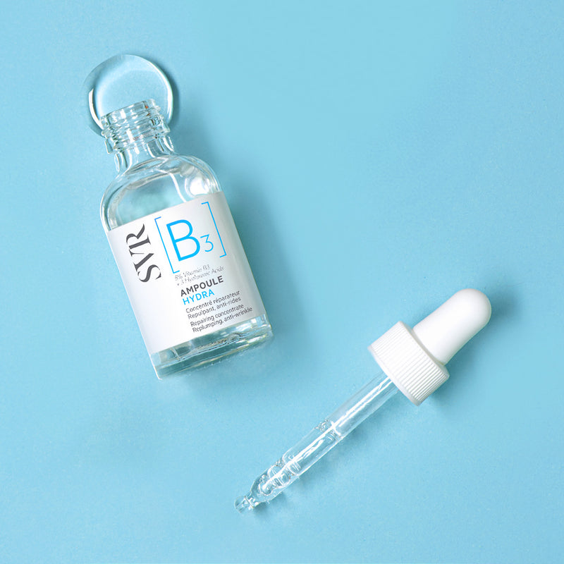 Flacon en verre transparent SVR Ampoule Hydra avec pipette blanche sur fond bleu clair.
