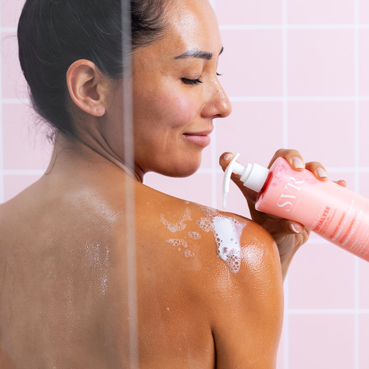 Femme appliquant le baume lavant SVR Topialyse sur son épaule dans une douche aux carreaux roses.