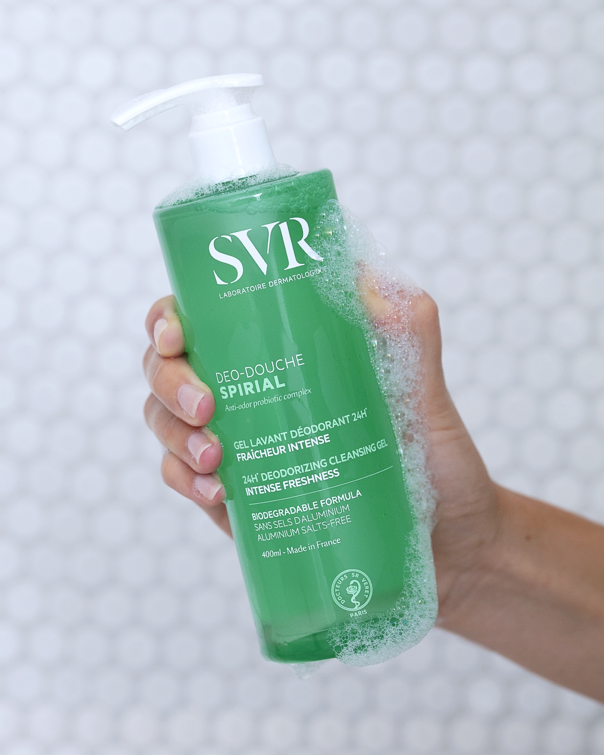 Flacon pompe vert de gel douche SVR Spirial 400ml tenu en main avec de la mousse, sur fond de carrelage blanc.
