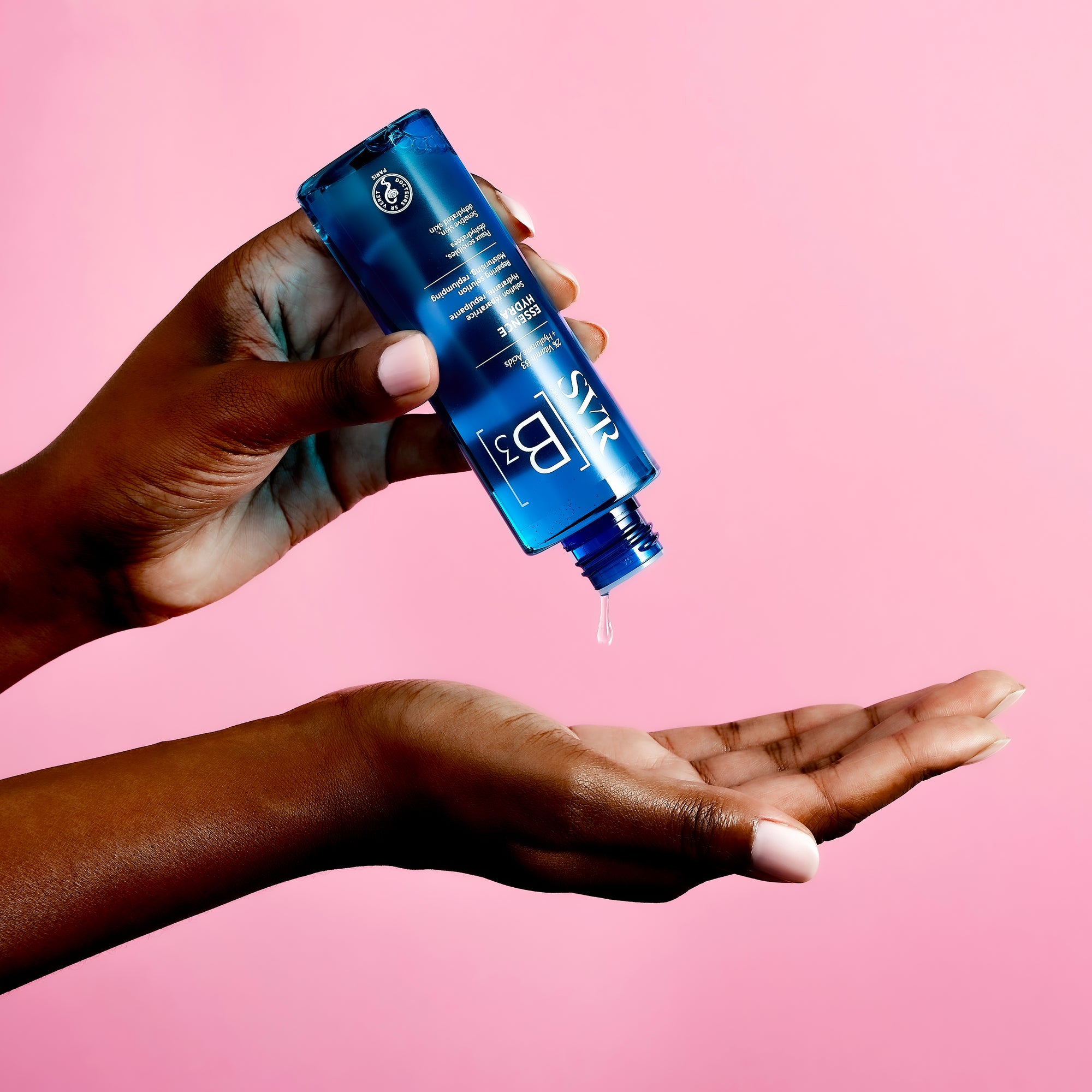 Flacon bleu SVR B3 Essence Hydra versant une goutte de soin hydratant dans une main sur fond rose.