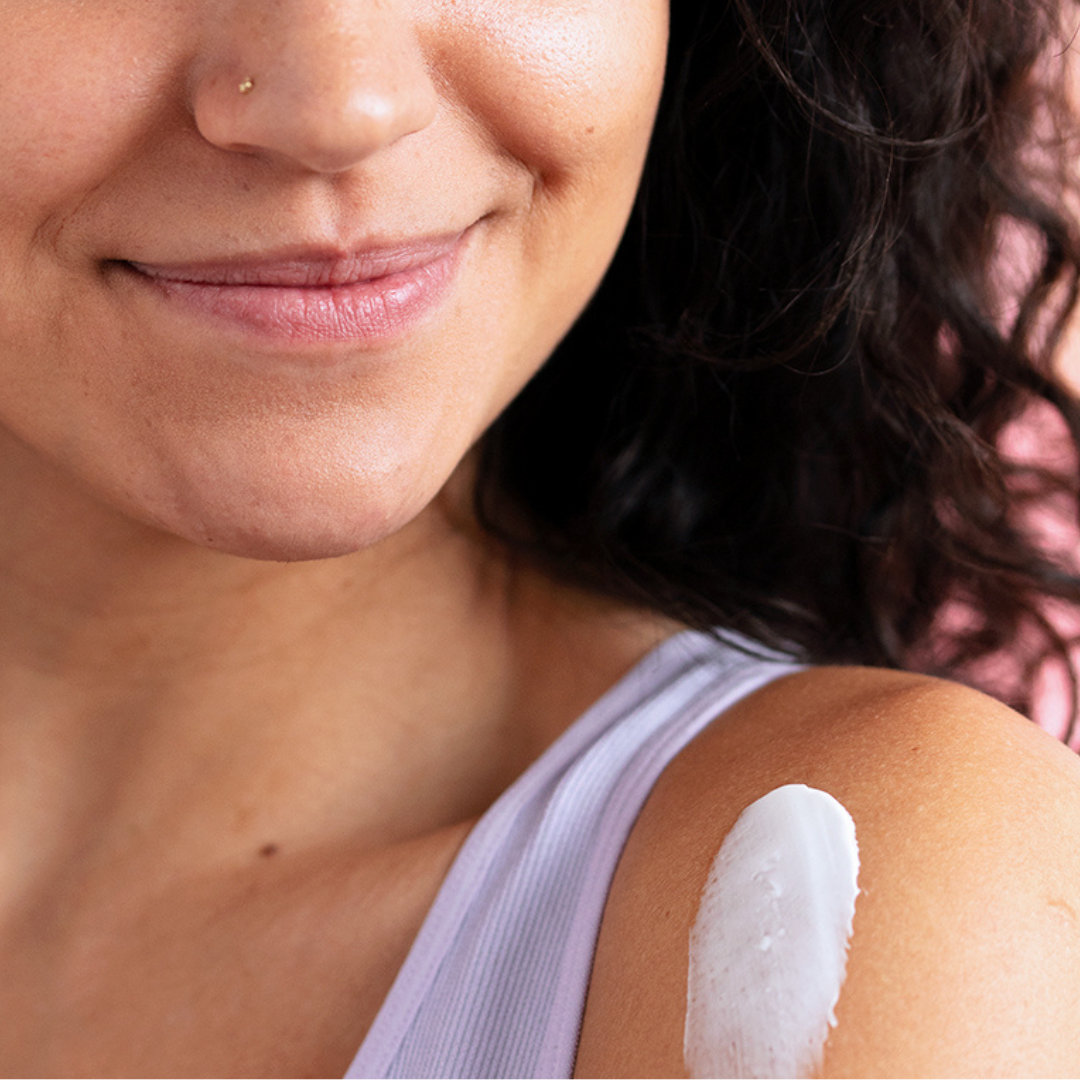 Femme souriante appliquant une noisette de crème hydratante blanche et onctueuse sur son épaule nue.