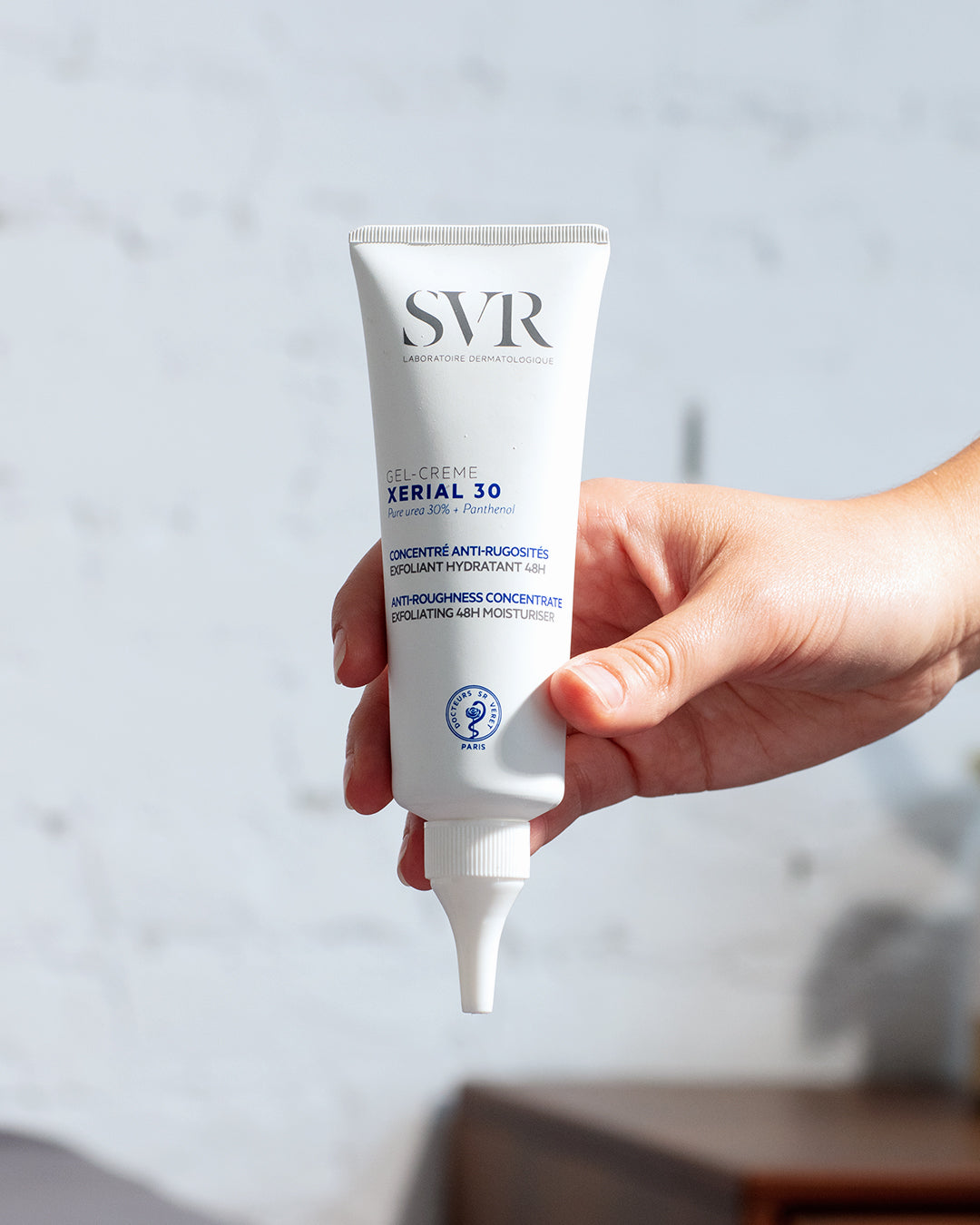 Tube blanc de gel-crème SVR XERIAL 30 avec embout applicateur précis, tenu dans une main sur fond texturé clair.