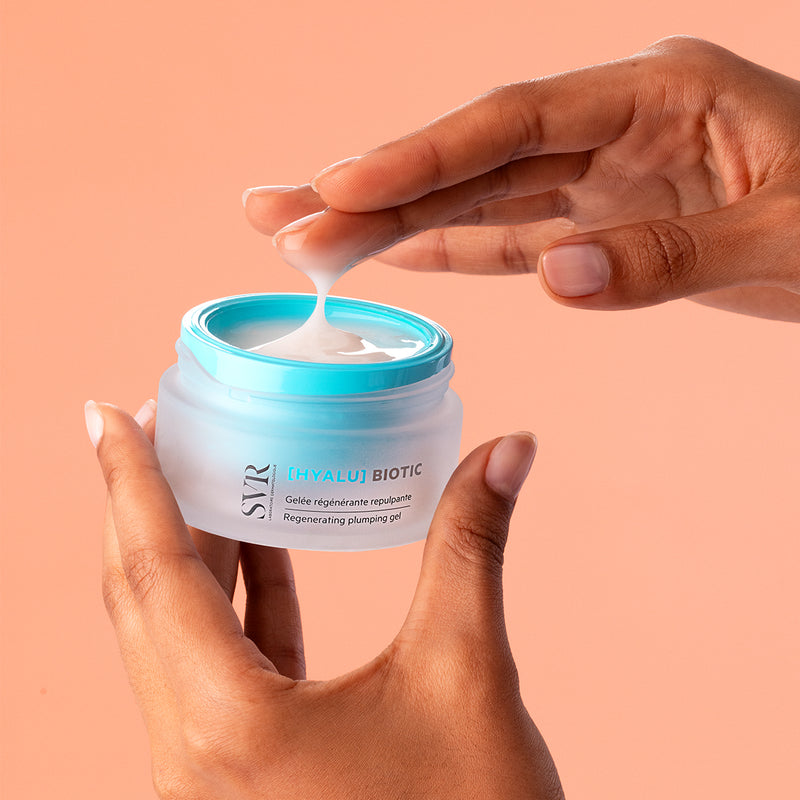 Hands holding a frosted glass jar of SVR Hyalu Biotic regenerating plumping gel with a bright blue lid and white texture.