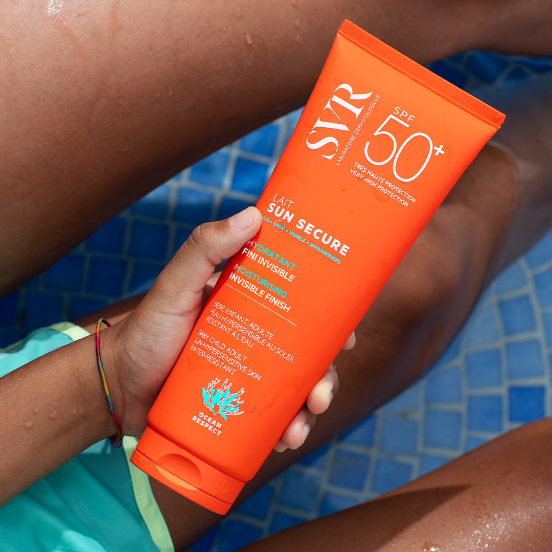 Tube orange de lait solaire SVR Sun Secure SPF50+ tenu par une main au-dessus d'une piscine bleue.