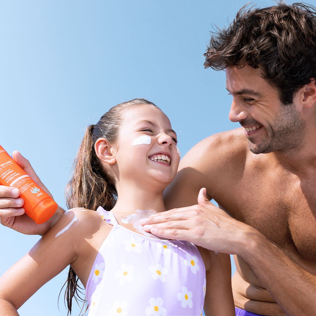Père appliquant du lait solaire SVR Sun Secure SPF50+ sur l'épaule de sa fille souriante sous un ciel bleu.