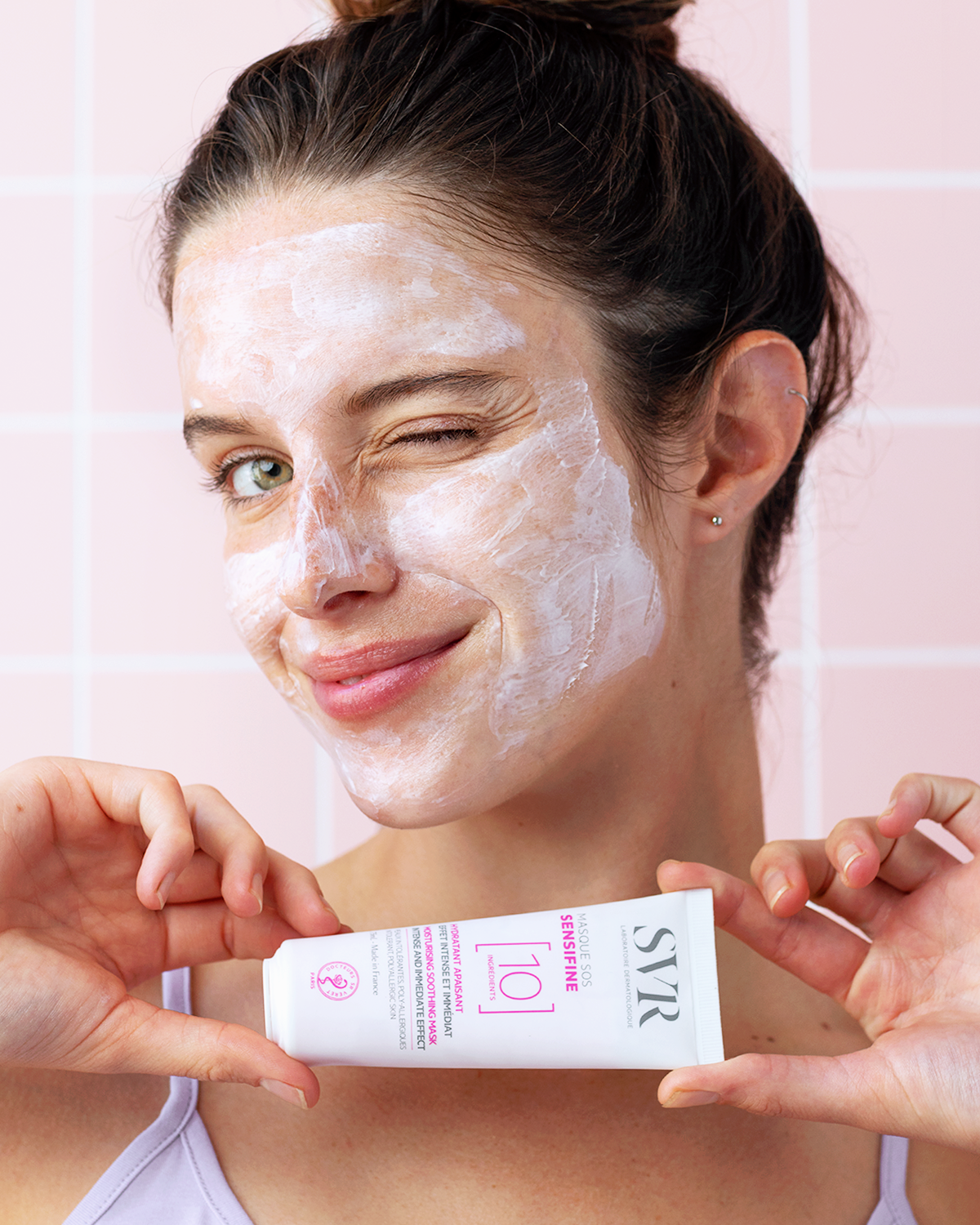 Femme souriante avec un masque visage blanc SVR Sensifine SOS et le tube du produit en main devant un carrelage rose.