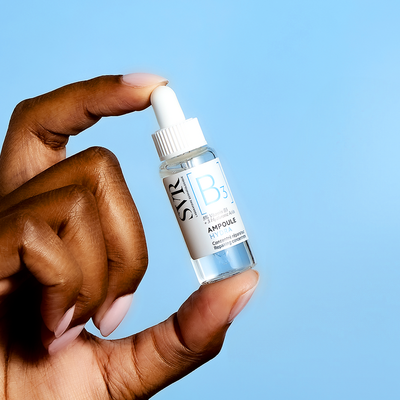 Main tenue par une main, flacon compte-gouttes transparent SVR Ampoule Hydra B3 sur fond bleu clair.