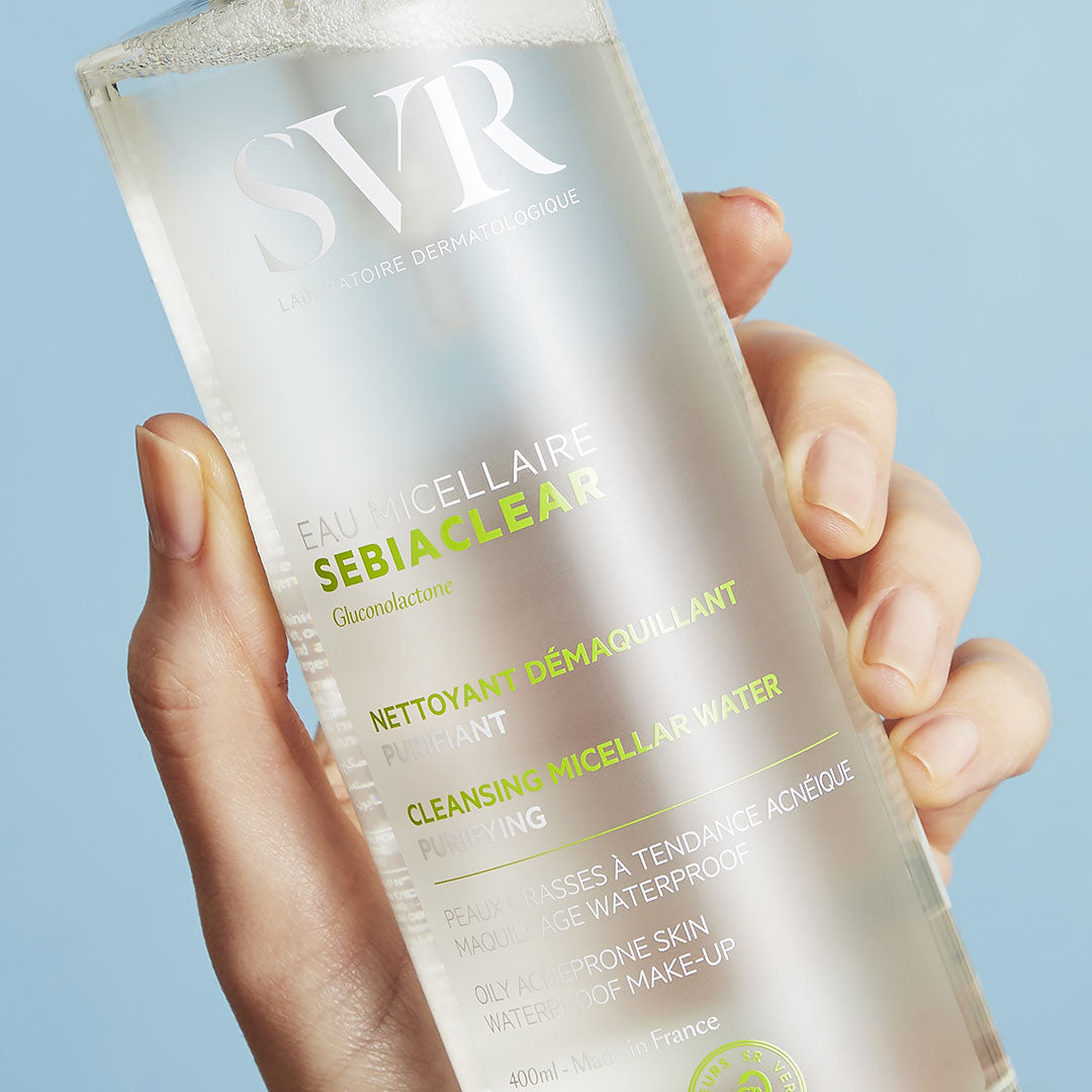 Flacon transparent de 400ml d'eau micellaire SVR Sebiaclear tenu en main sur fond bleu clair.