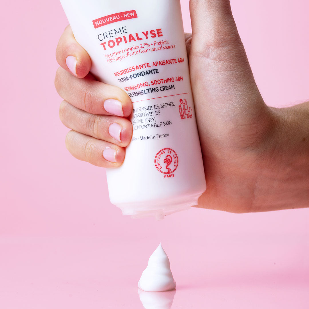Main tenue en main pressant un tube blanc de crème hydratante Topialyse sur fond rose, avec une noisette de produit onctueux.