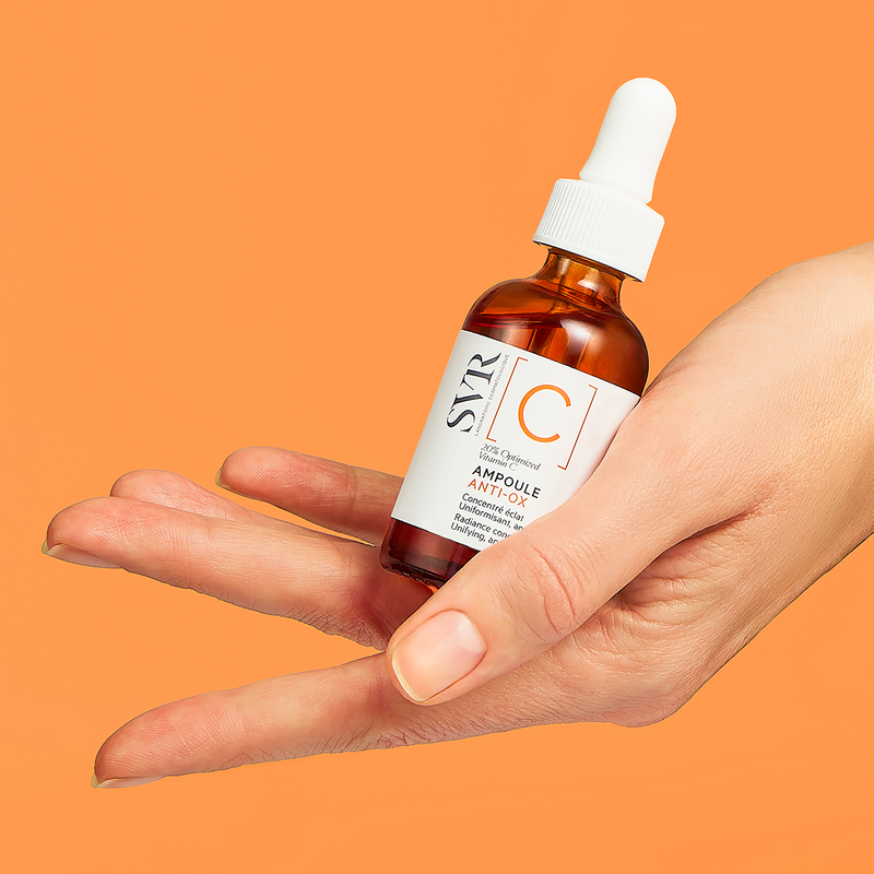 Main tenue en main d'un flacon compte-gouttes SVR Ampoule Anti-Ox à la vitamine C sur fond orange vif.