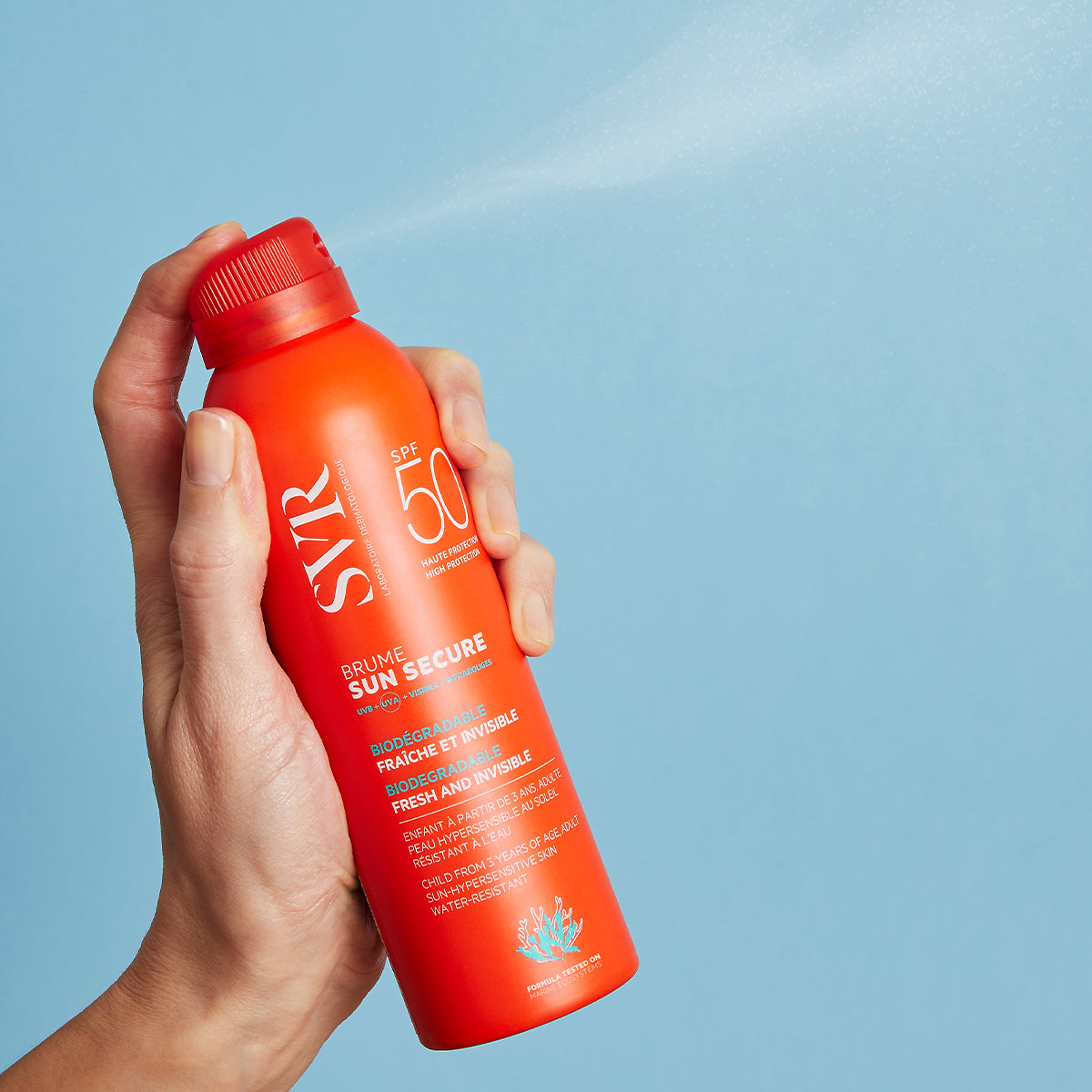 Main tenant un spray solaire orange SVR SUN SECURE SPF50 diffusant une brume légère sur fond bleu.