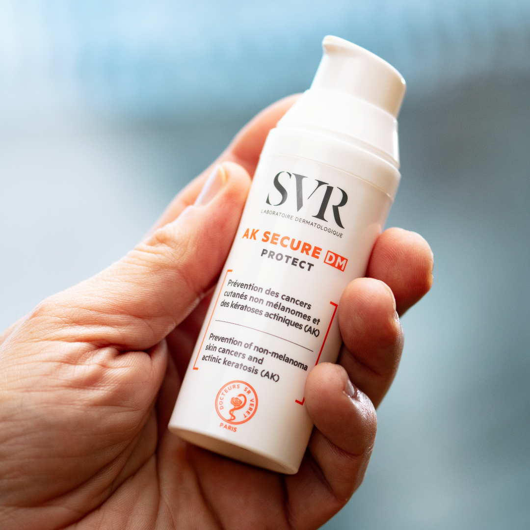 Flacon pompe blanc SVR AK SECURE DM Protect tenu en main, soin dermatologique pour la prévention des kératoses actiniques.