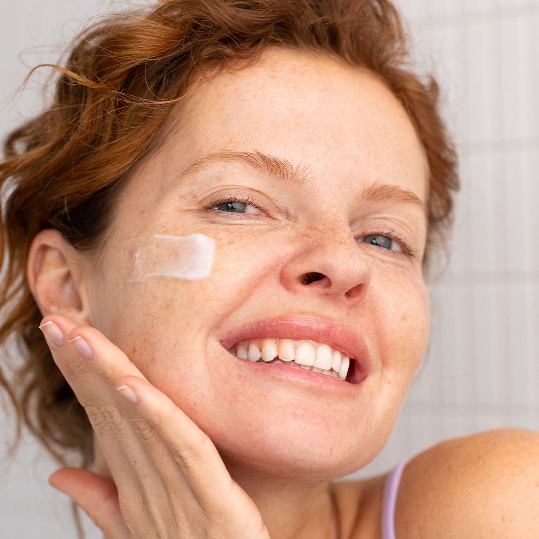 Femme rousse souriante appliquant une crème régénérante blanche sur sa joue pour lisser et matifier sa peau sensible.