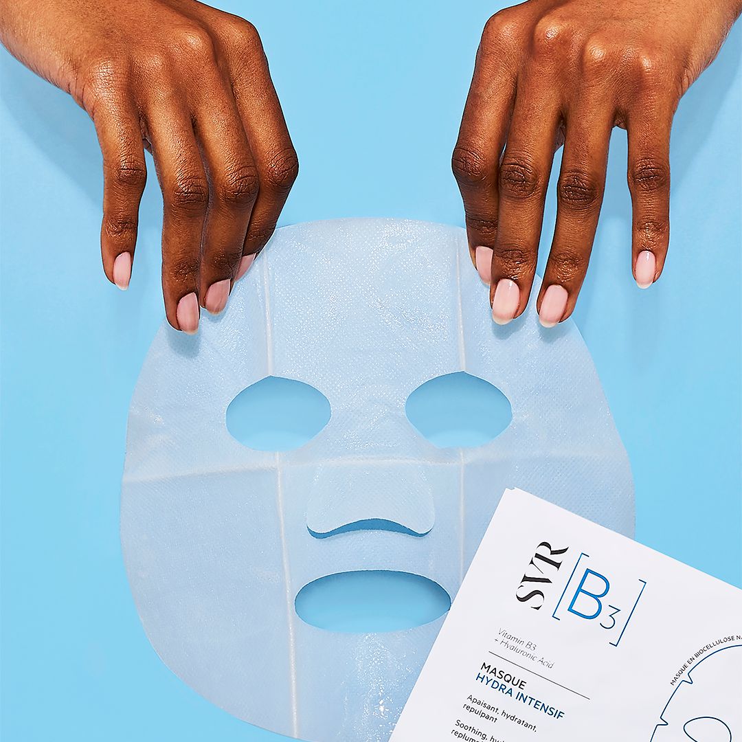 Mains tenant un masque en biocellulose bleu clair SVR B3 Hydra Intensif sur fond bleu.