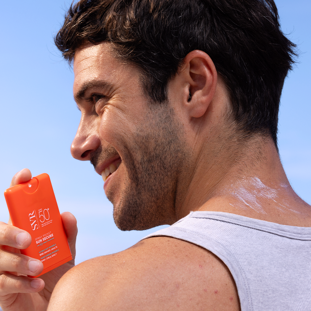 Homme souriant tenant un flacon spray SVR Sun Secure SPF50+ orange format poche contre un ciel bleu ensoleillé.