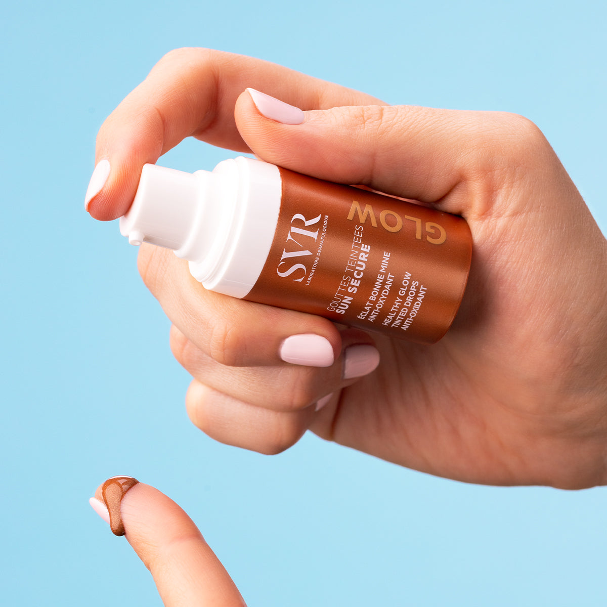 Hand holding a bronze SVR Sun Secure tinted drops bottle, dispensing a drop of brown liquid onto a fingertip against blue.