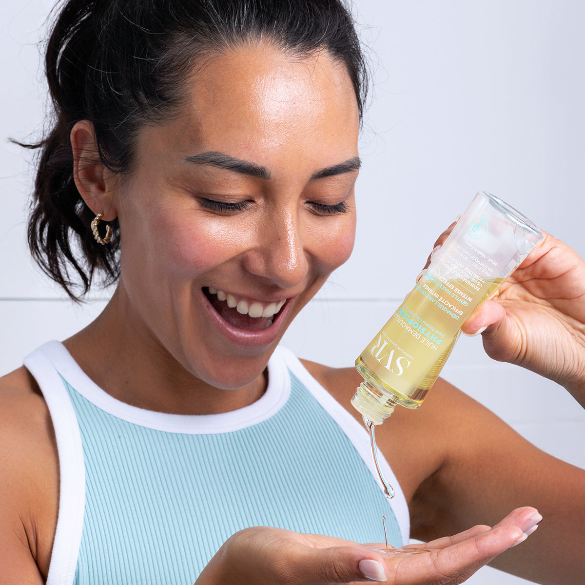 Femme souriante versant l'huile démaquillante SVR Physiopure dans sa main, flacon transparent et texture fluide dorée.