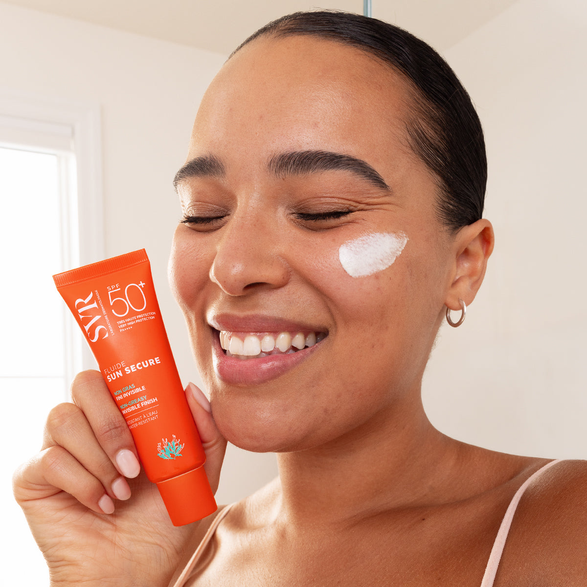 Femme souriante tenant un tube orange de fluide solaire SVR SUN SECURE SPF50+ avec une touche de crème sur la joue.