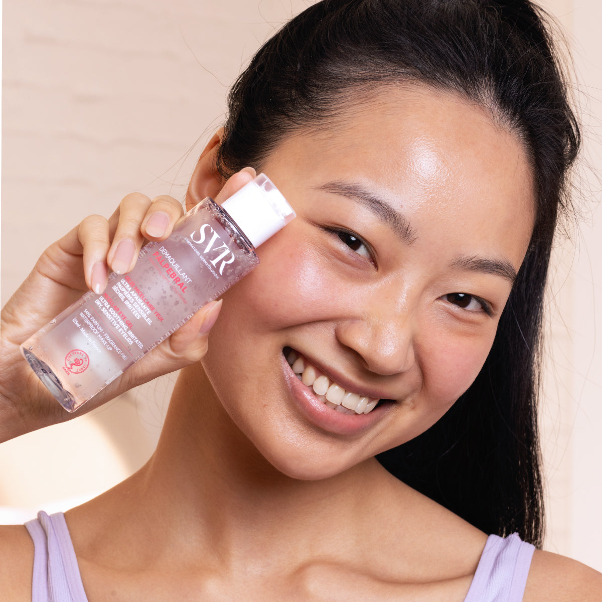 Femme souriante tenant un flacon transparent de gel démaquillant SVR Palpebral de 125 ml avec bouchon blanc.