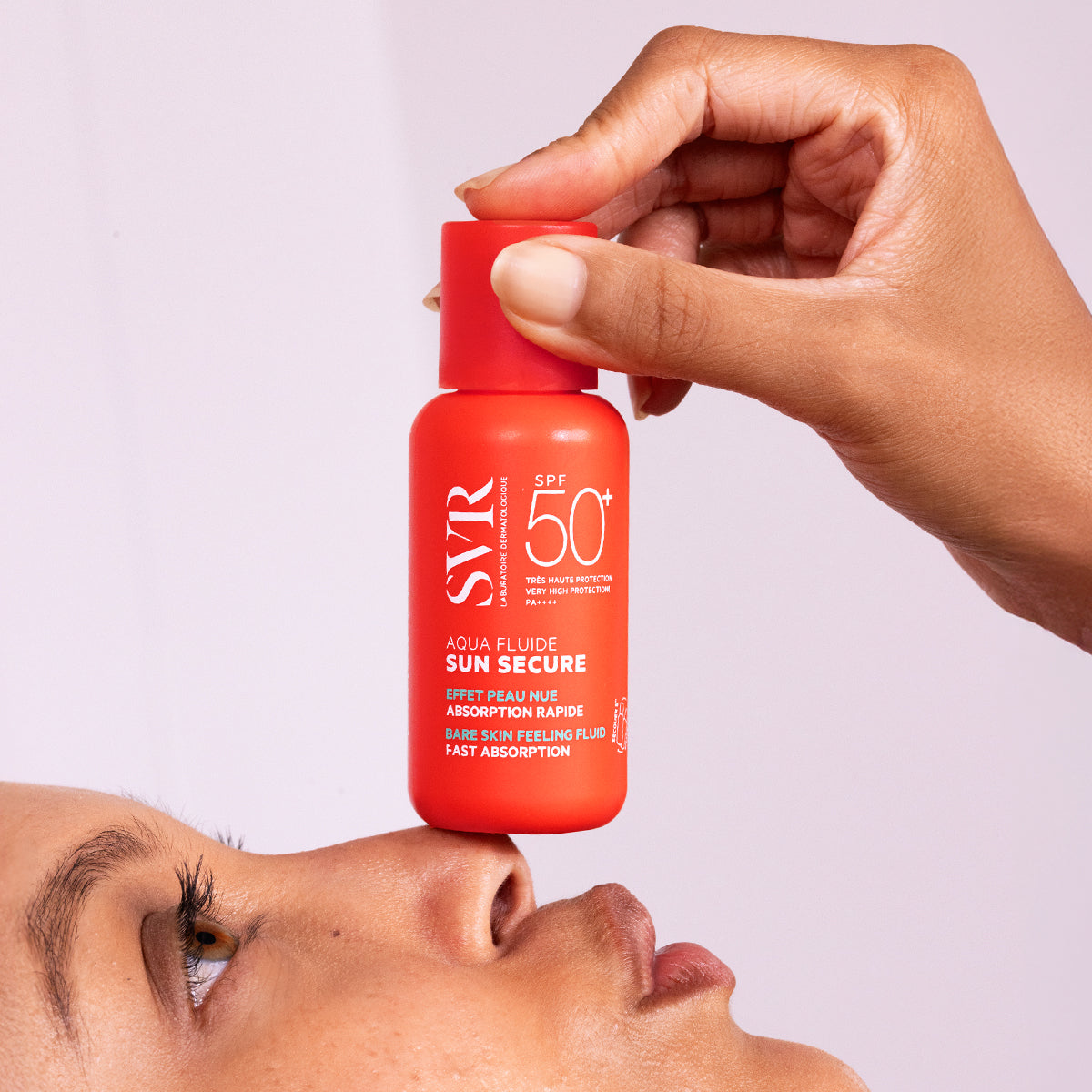 Hand holding a bright orange SVR SUN SECURE Aqua Fluide SPF50+ bottle above a person's nose against a light background.