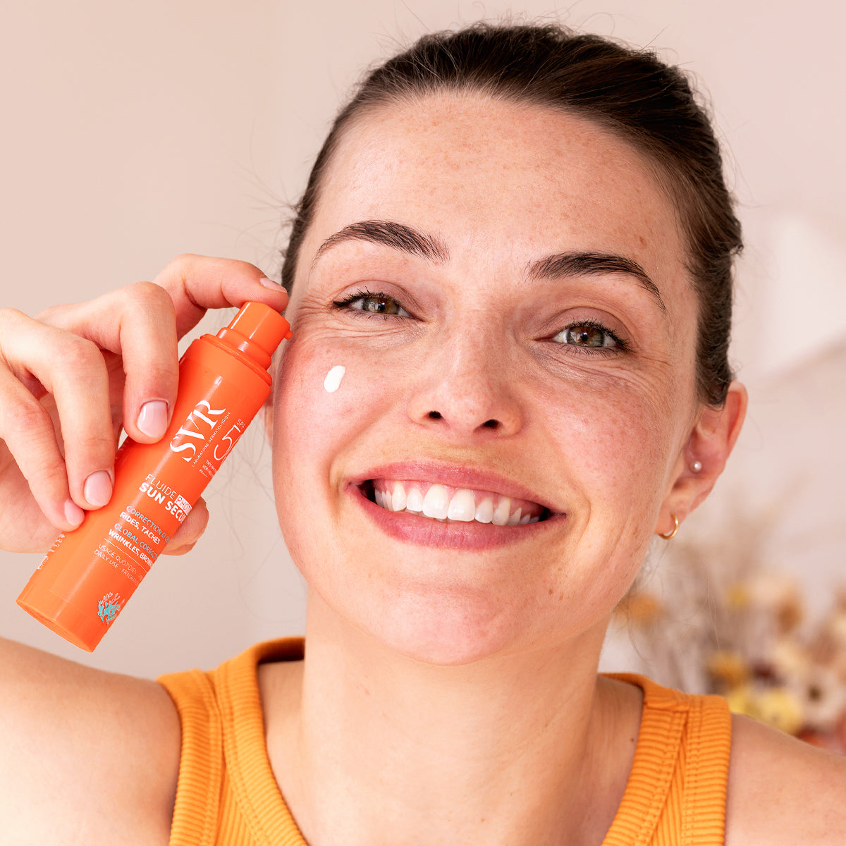 Femme souriante tenant un flacon pompe orange SVR Sun Secure Fluide Photo-Age SPF50+ près de son visage.