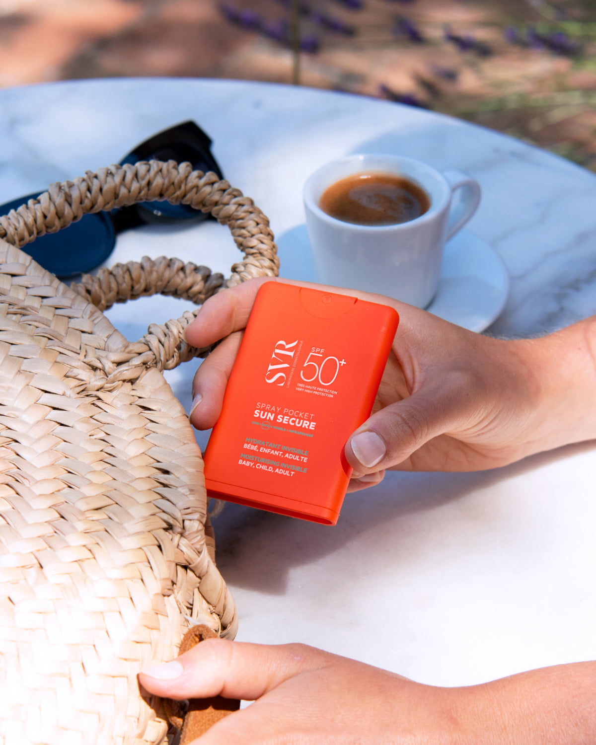 Main orange SVR SUN SECURE SPF50+ pocket spray tenu en main près d'un sac en paille et d'une tasse de café.