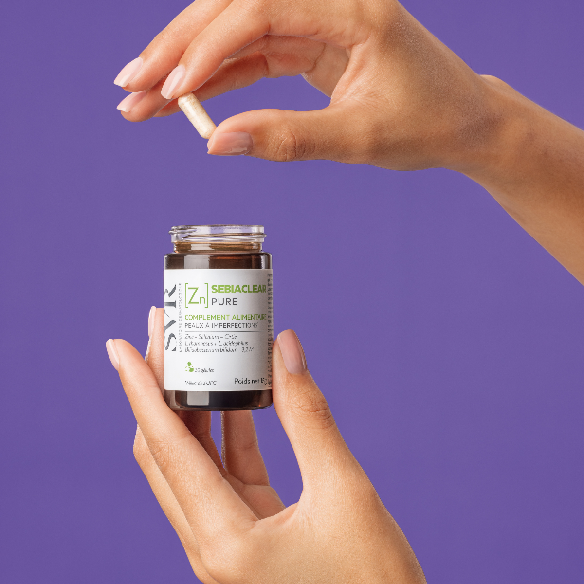 Hands holding a small brown glass bottle of SVR Sebiaclear Pure supplement capsules against a purple background.