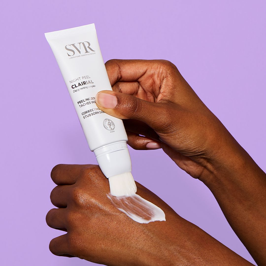 Tube blanc de soin SVR Clairial Night Peel avec applicateur pinceau appliquant une crème sur le dos d'une main.