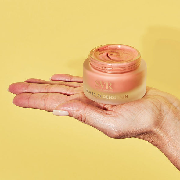 Pot en verre dépoli de crème SVR Densitium Rose Éclat à la texture rosée, tenu dans une main sur fond jaune.