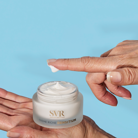 Pot en verre dépoli SVR DENSITIUM Crème Riche avec texture onctueuse blanche, tenu dans une main sur fond bleu.