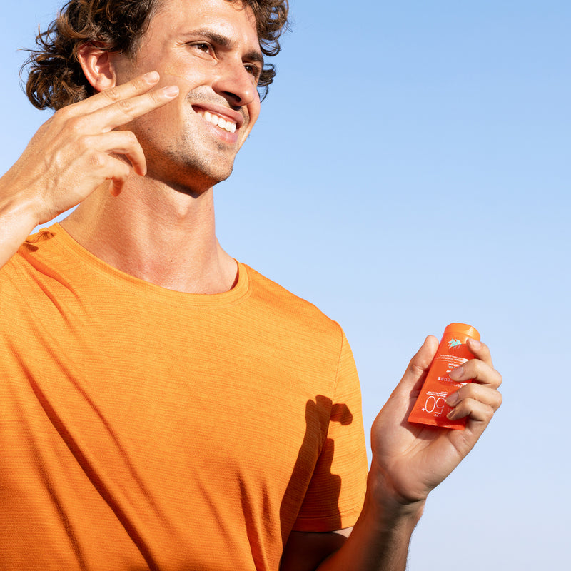 Smiling man in an orange shirt applying SPF 50+ sunscreen to his face against a clear blue sky.