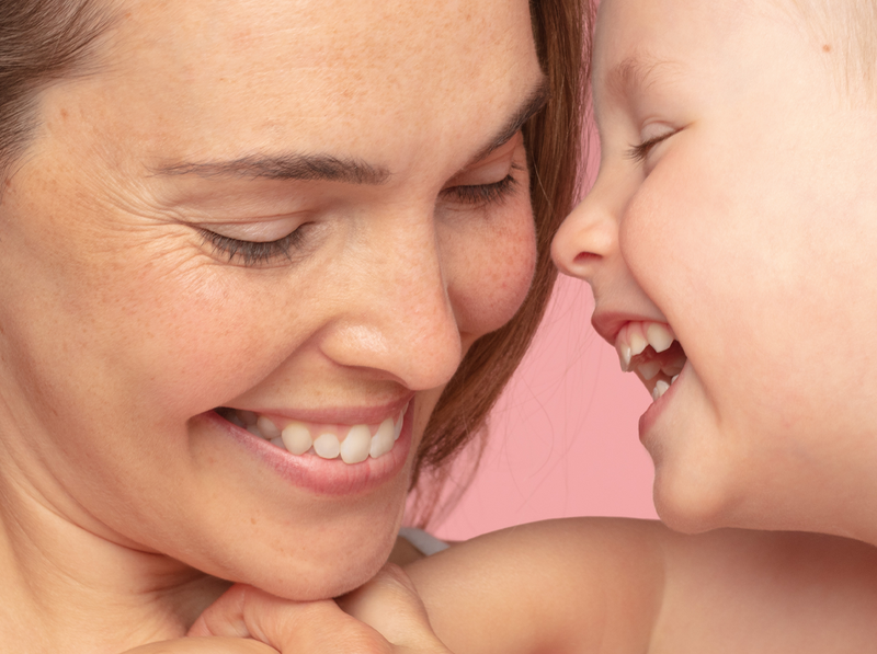 Mère et son jeune enfant souriants, peau saine et lumineuse sur fond rose, évoquant la douceur et le soin.