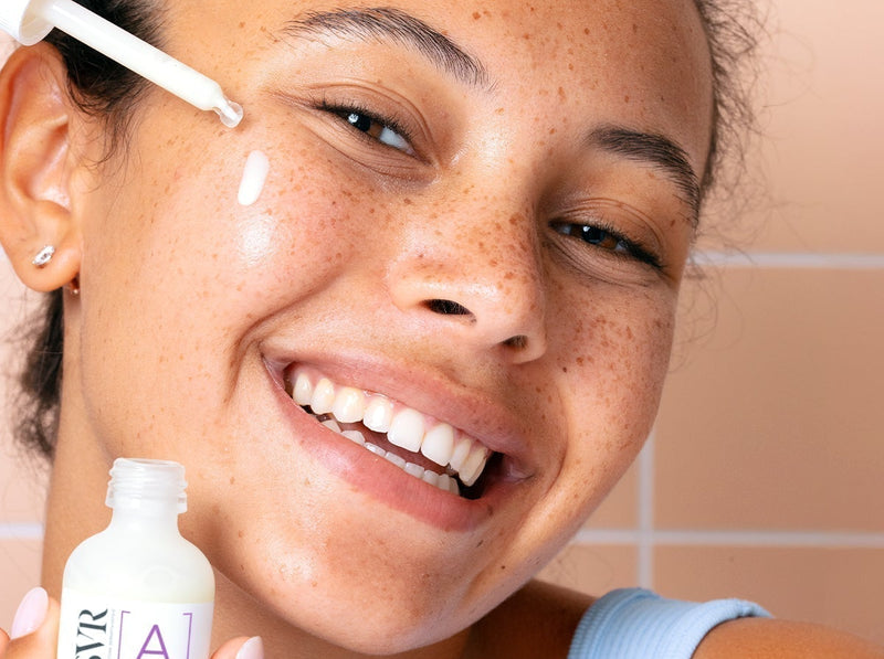 Femme souriante appliquant un sérum au rétinol SVR sur son visage devant un carrelage rose.