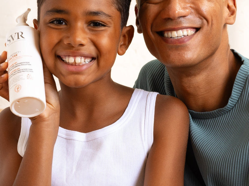 Un père et son fils souriants, le garçon tenant un flacon de crème hydratante SVR avec une touche de produit sur le bras.