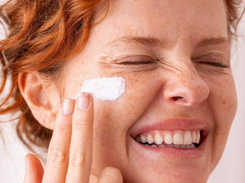 Femme rousse souriante appliquant une crème hydratante blanche sur sa joue pour une routine soin anti-âge.