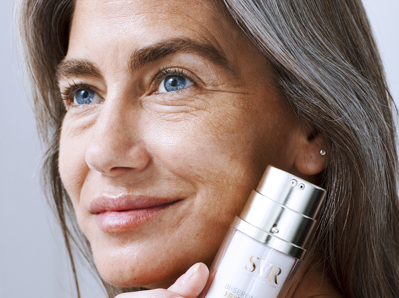 Femme mature aux cheveux gris tenant un flacon de sérum anti-âge SVR pour le soin du visage.