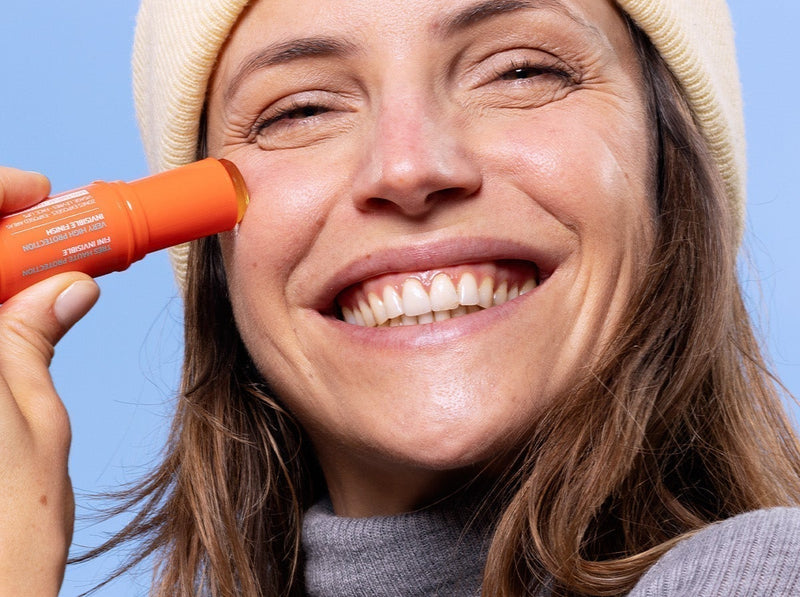 Femme souriante en bonnet et pull gris appliquant un stick de crème solaire orange sur sa joue sous un ciel bleu.