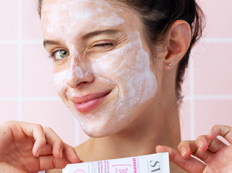 Jeune femme souriante avec un masque apaisant blanc sur le visage tenant un tube de soin SVR Sensifine.