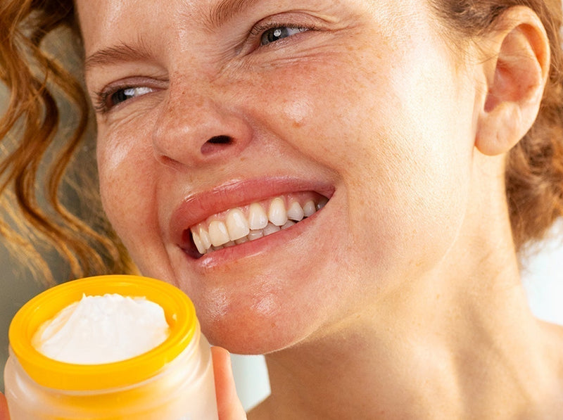 Femme rousse souriante tenant un pot de crème anti-âge SVR au couvercle jaune vif.