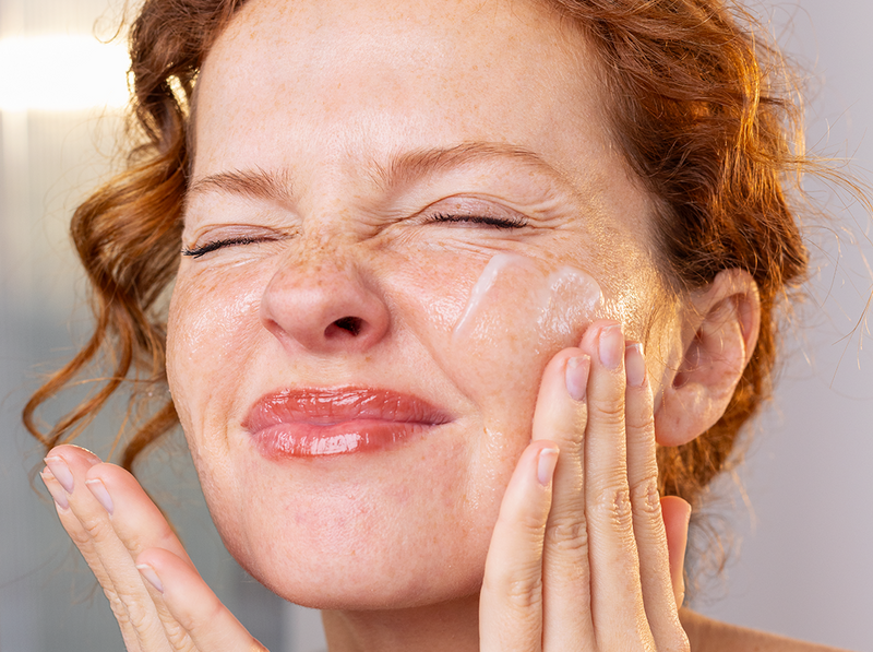 Femme rousse aux taches de rousseur appliquant une crème hydratante apaisante sur son visage pour soulager sa peau.