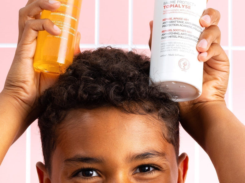 Enfant souriant tenant deux flacons de produits de soin SVR Topialyse devant un carrelage rose.