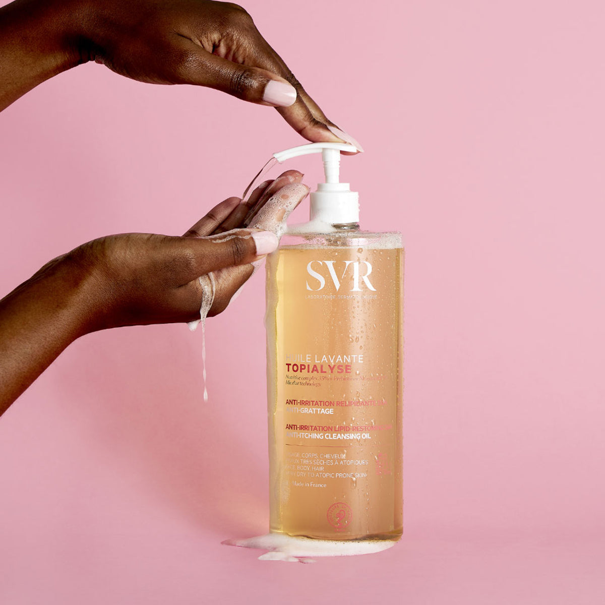 Flacon pompe transparent de SVR Topialyse Huile Lavante avec texture huile dorée, utilisé par des mains sur fond rose.