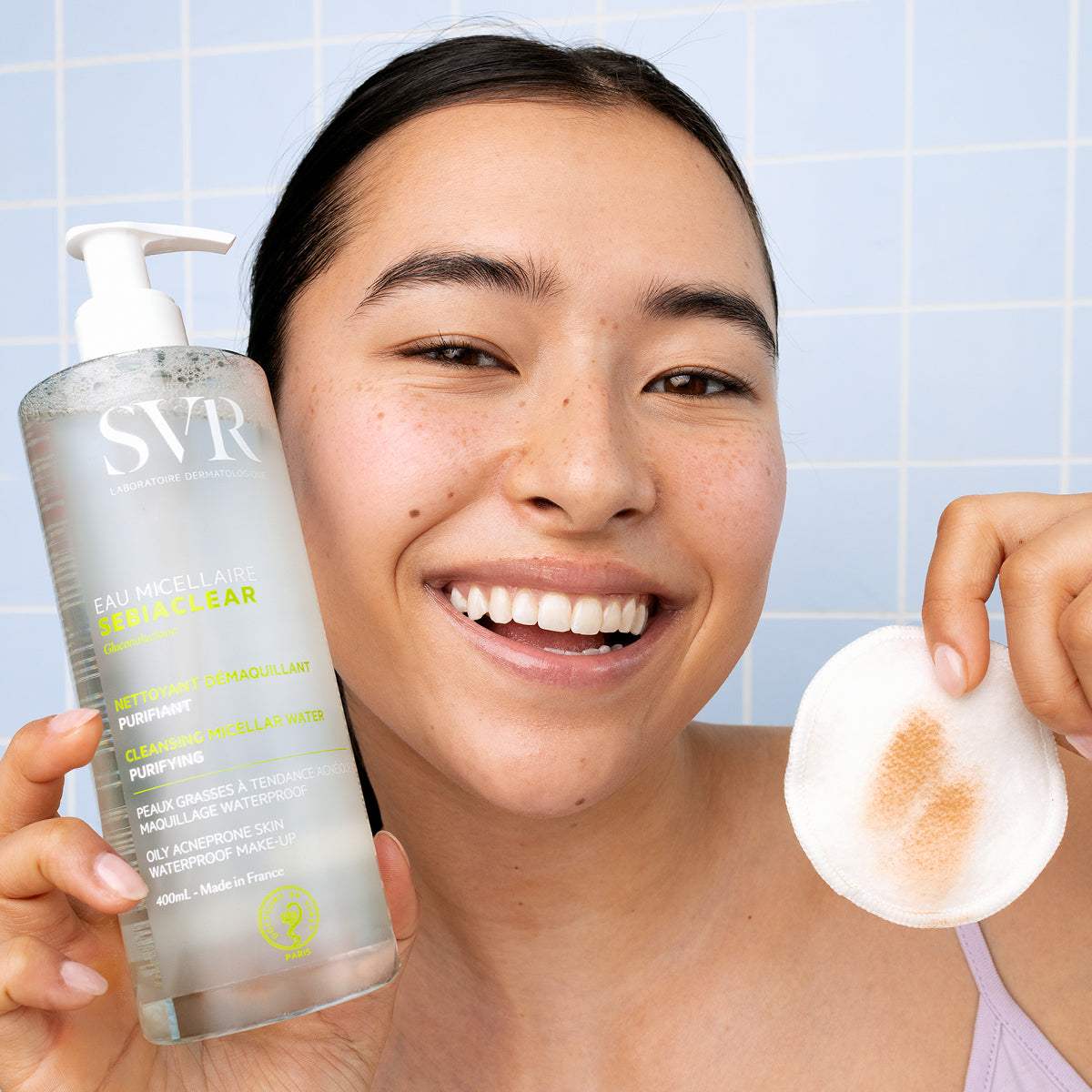 Femme souriante tenant un flacon pompe SVR Sebiaclear Eau Micellaire de 400ml et un coton démaquillant usagé.