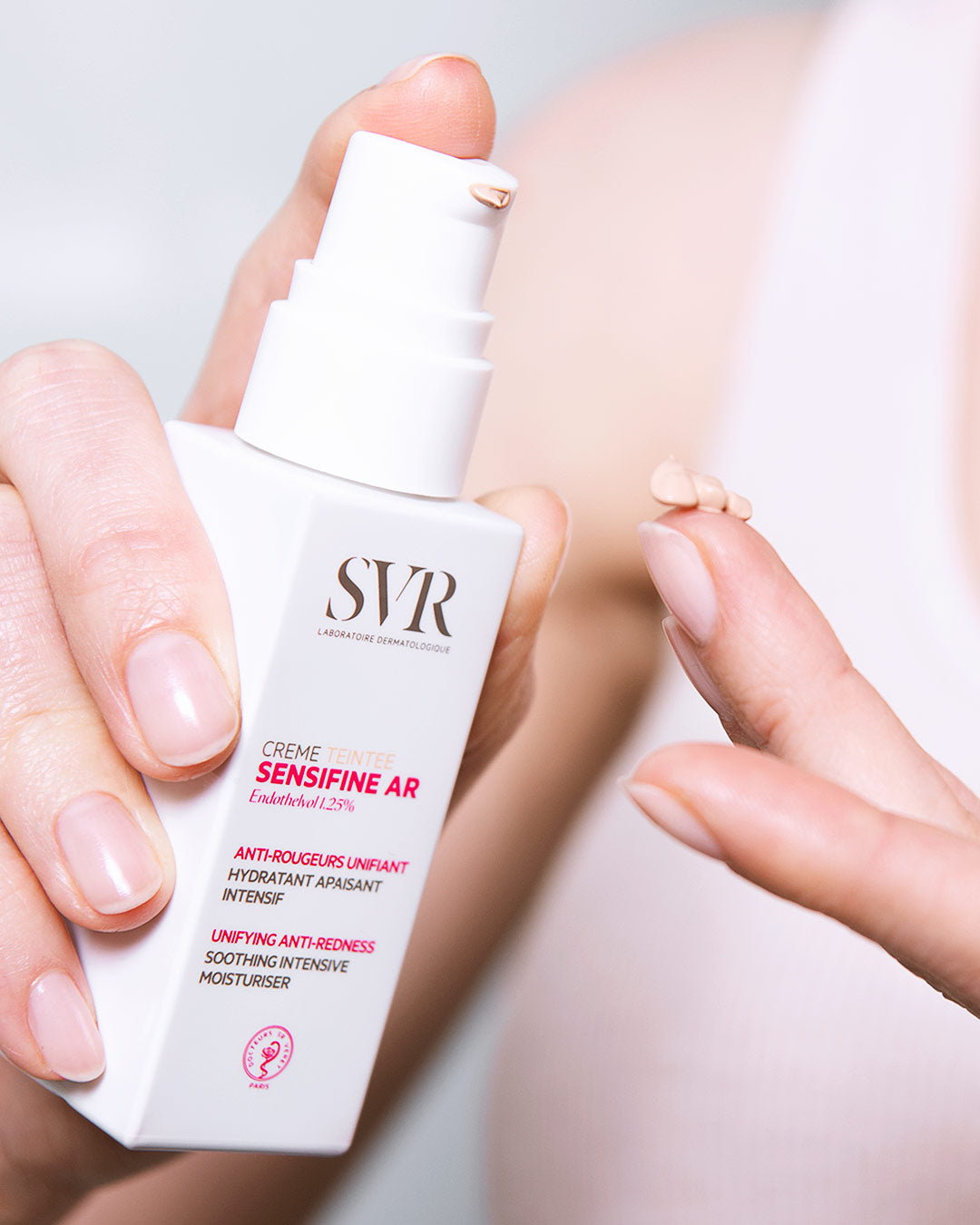 Flacon pompe blanc SVR Sensifine AR Crème Teintée avec une noisette de crème beige sur le doigt pour corriger les rougeurs.