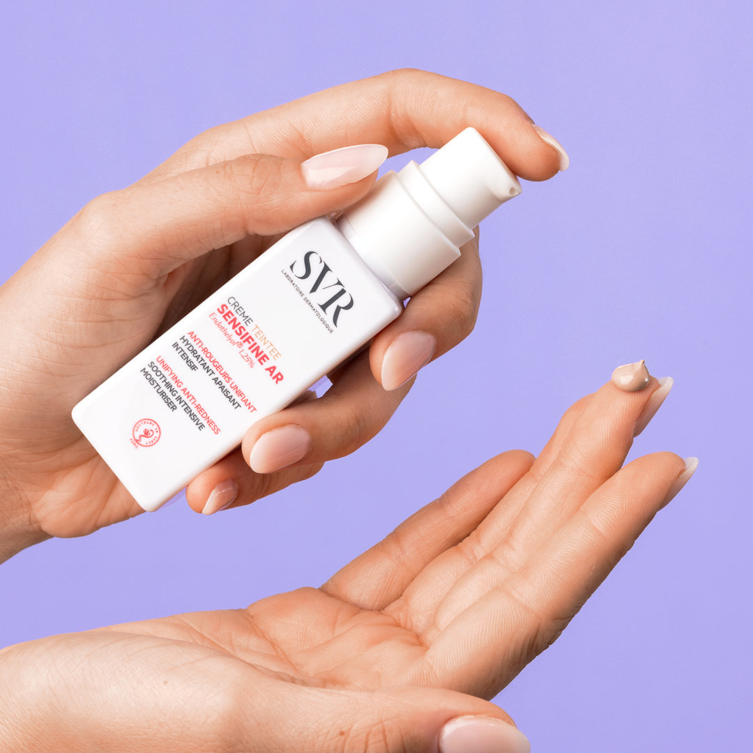 Hand holding a white SVR Sensifine AR Crème Teintée pump bottle with a small drop of tinted cream on a fingertip.