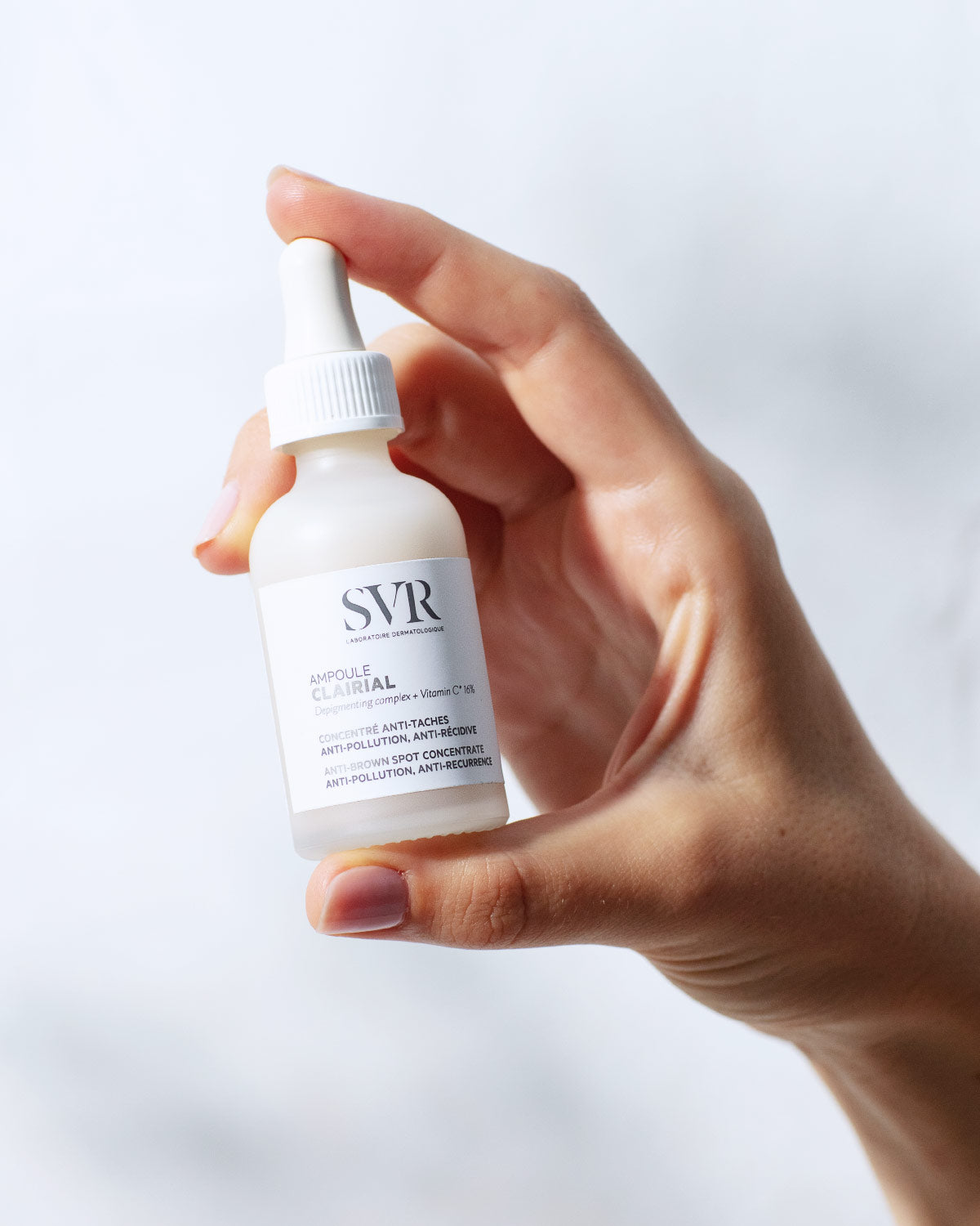 Main tenue en main d'un flacon compte-gouttes SVR Clairial Ampoule blanc et givré contre un fond clair.