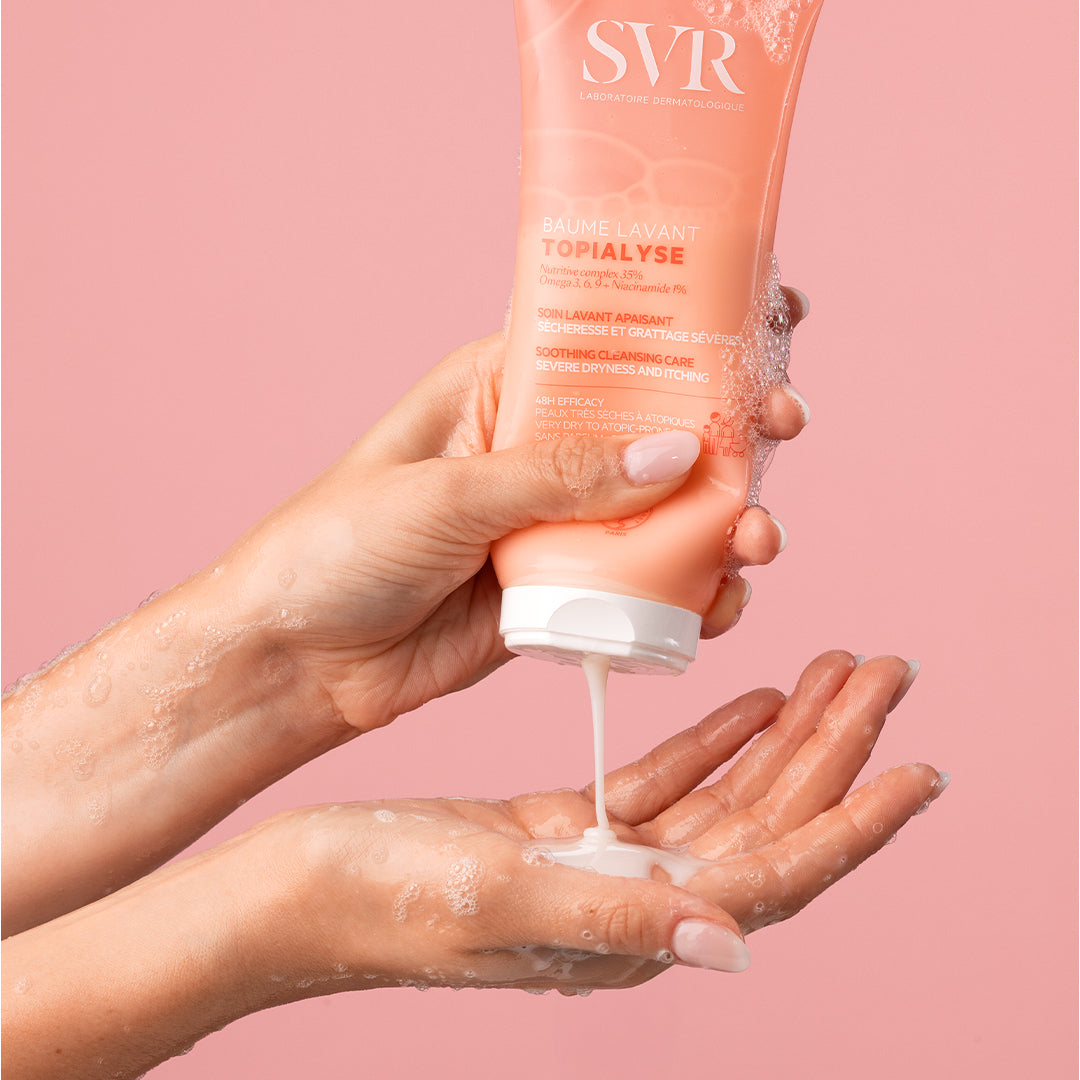 Tube de SVR Topialyse Baume Lavant rose pressé dans les mains, délivrant une texture crémeuse sur fond rose.