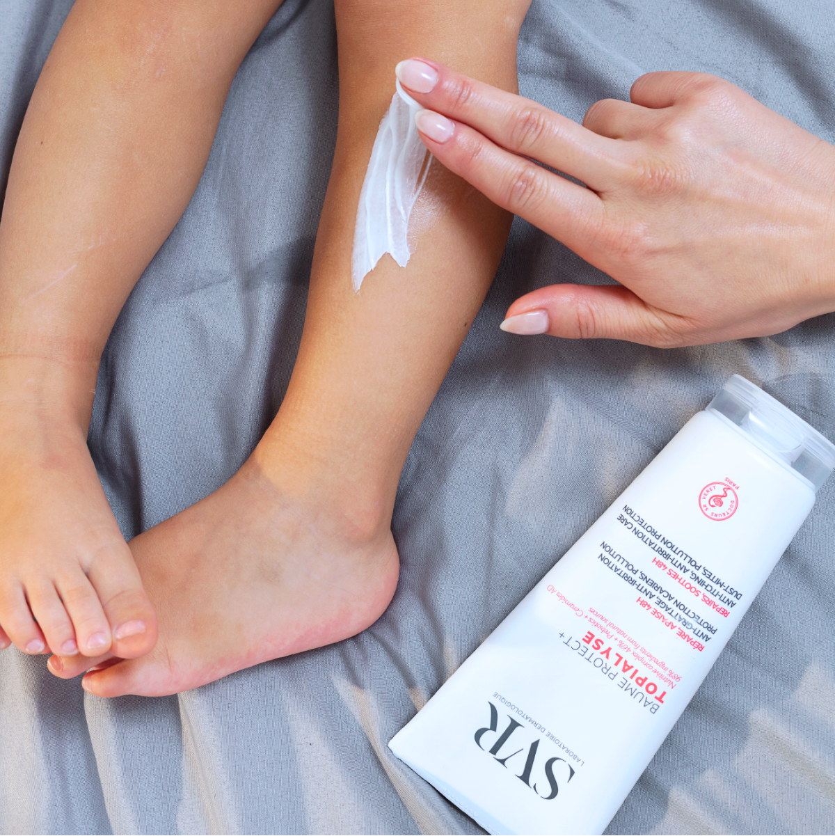 Main appliquée sur la jambe d'un enfant pour appliquer la crème SVR Topialyse sur un drap gris.